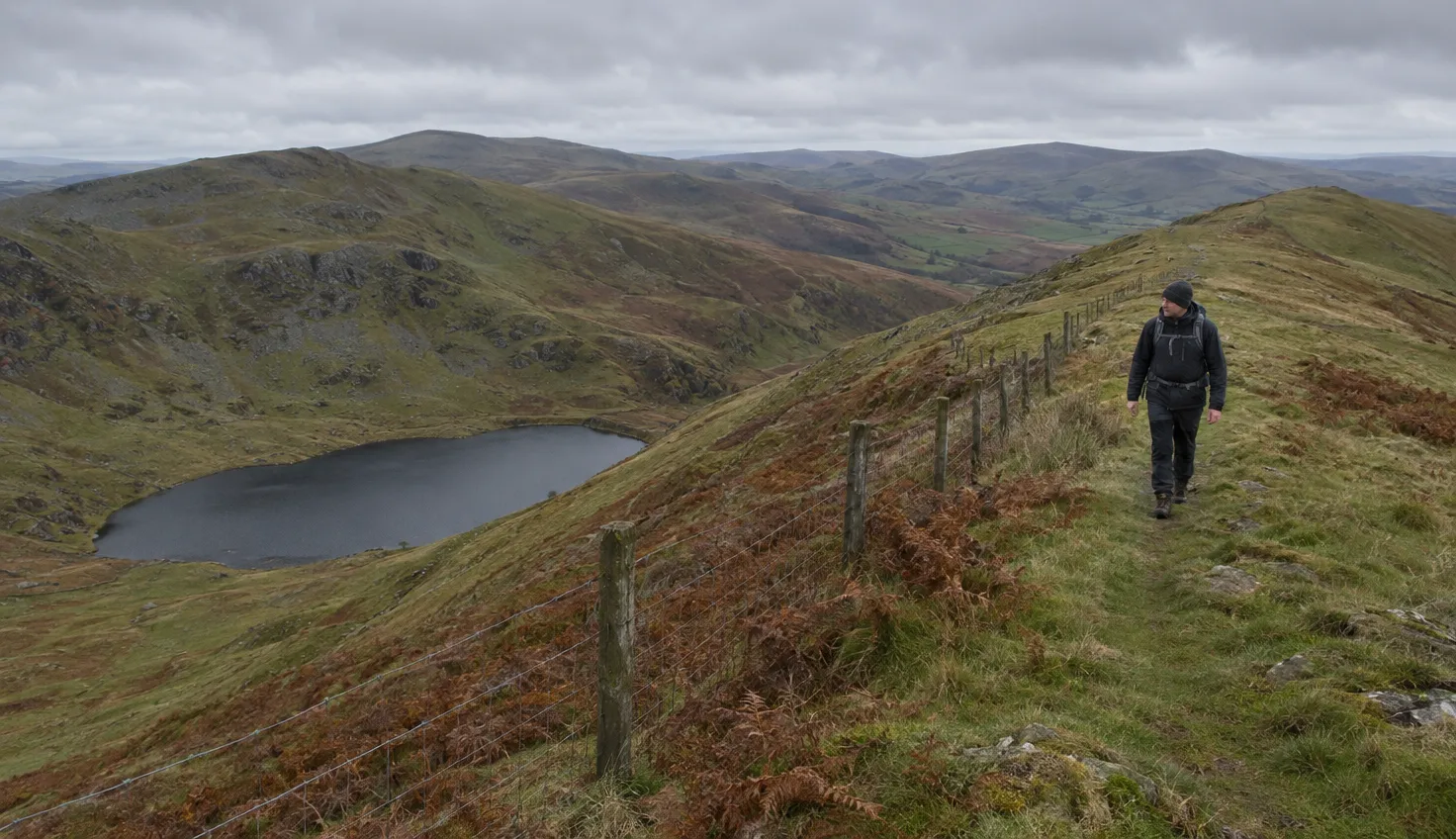 A single walker on a long grassy ridge beside a fence, a small dark tarn in a hollow below.