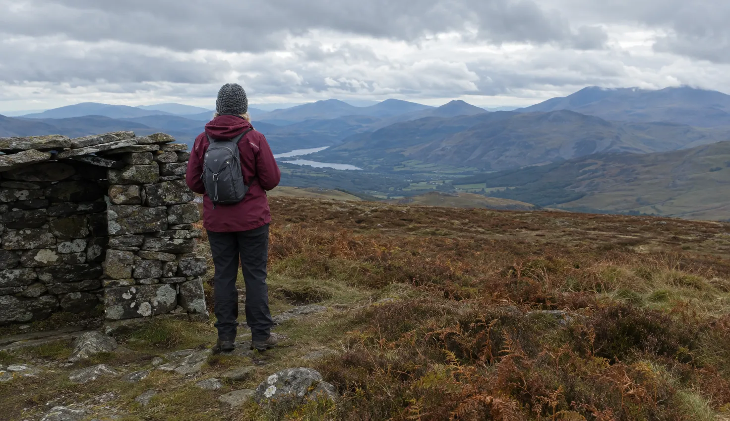 A walker on a heathery moorland summit beside a low stone shelter, looking west to distant mountains.