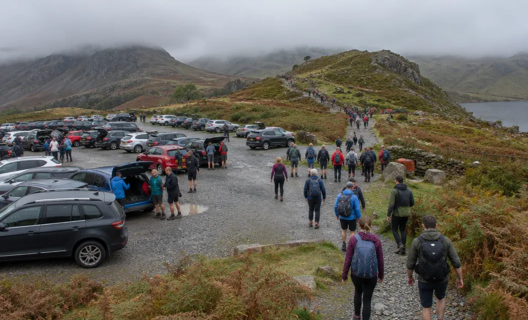 A crowded car park and path start with many walkers below cloudy hills.