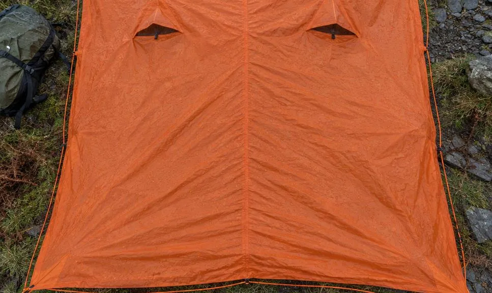 A group emergency shelter laid out flat and open on wet mountain grass, its full rectangular footprint visible from above with a walker's boots at the edge for scale.