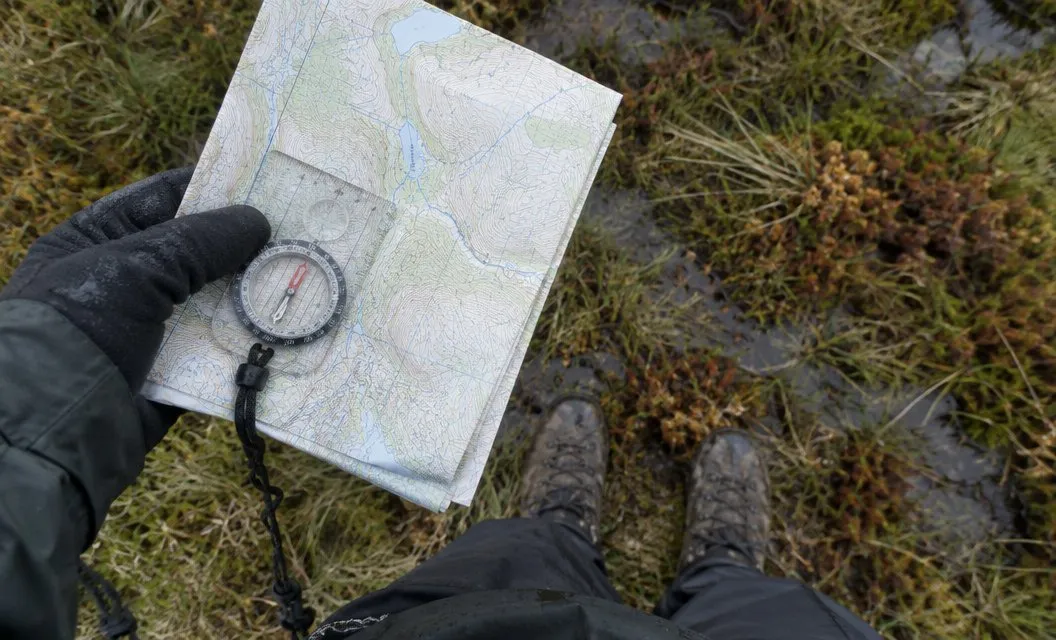 A gloved hand holding a compass on a folded map above muddy boots in boggy grass.