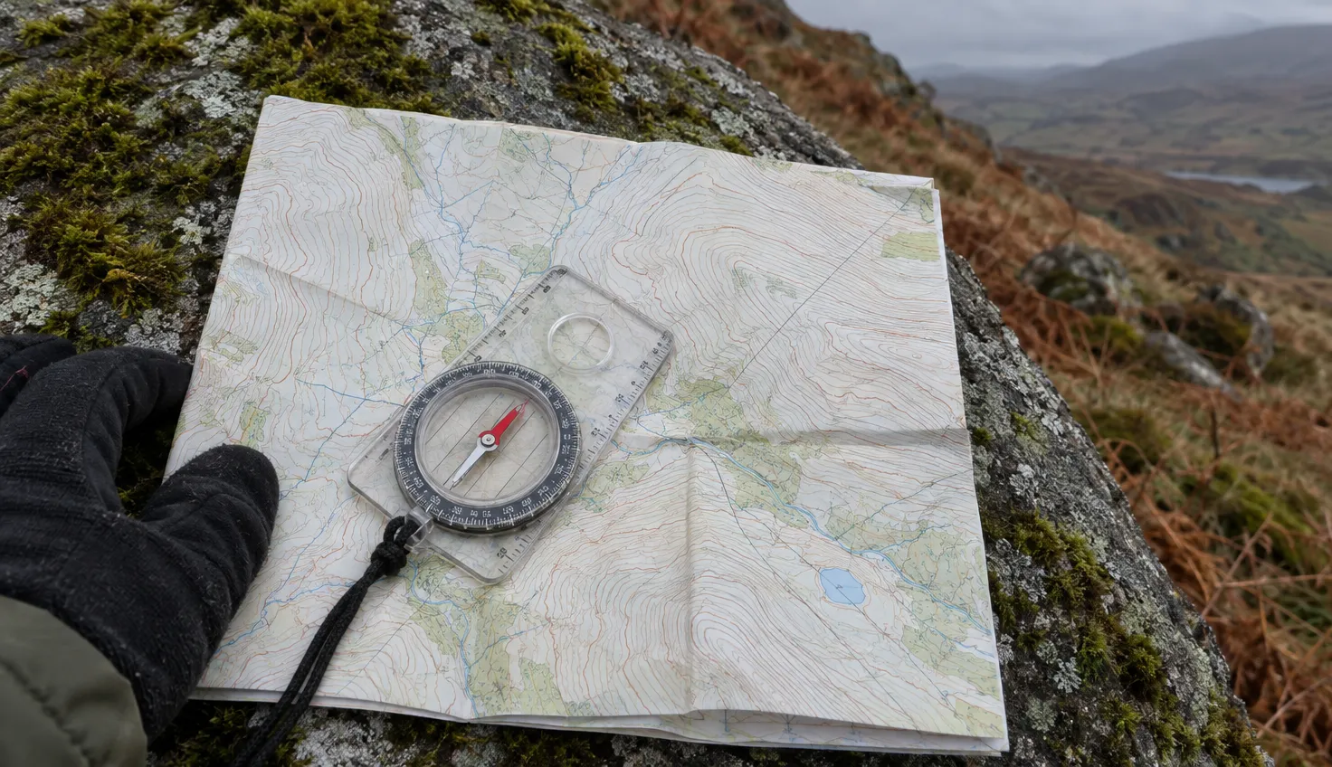 A clear baseplate compass lying on a folded paper map on a mossy boulder.