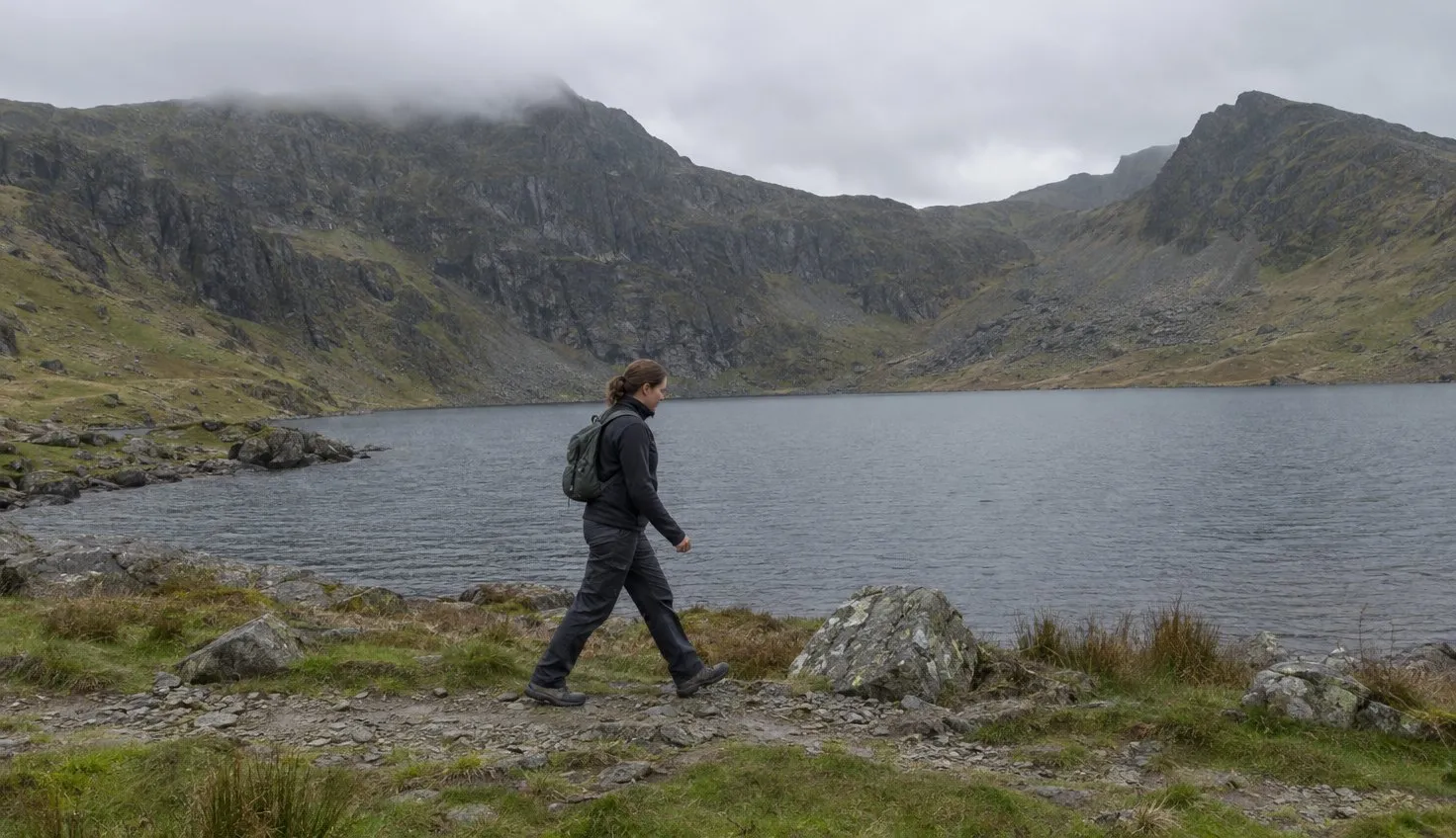 A lone walker on a stony lakeside path below steep dark cliffs, the lake flat and grey.