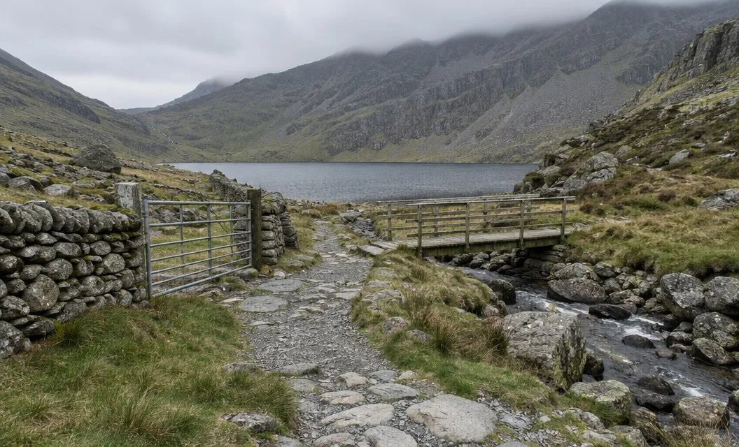 A stone path and a wooden footbridge leading towards a lake in a rocky mountain bowl.