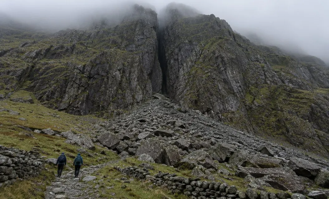 A dark cleft splitting a cliff face above a slope of boulders and scree, two small walkers below it.