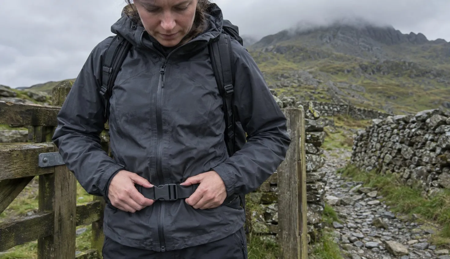 A walker at a wooden gate clipping the hip belt of a daypack before setting off up a mountain path.