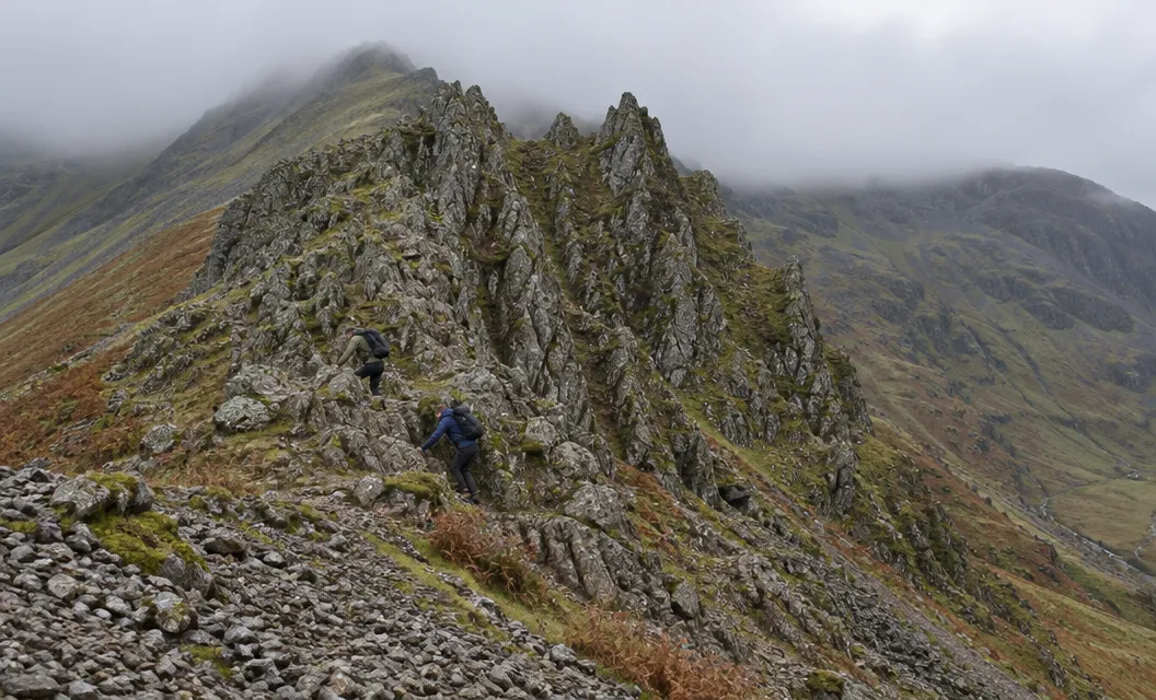 Two small walkers using their hands low on a steep ridge of jagged rock pinnacles above a scree slope.