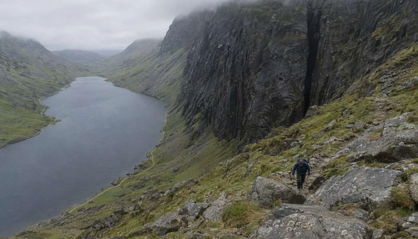 A walker on the boulder path beside the Devil's Kitchen cleft at the head of Cwm Idwal, with Llyn Idwal far below.