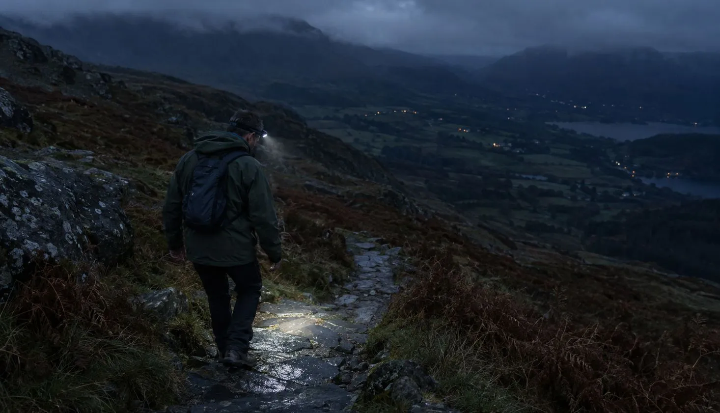 A lone walker descending wet slate steps on a Welsh mountain path after sunset, a head torch beam lighting the ground a few metres ahead and valley lights far below.