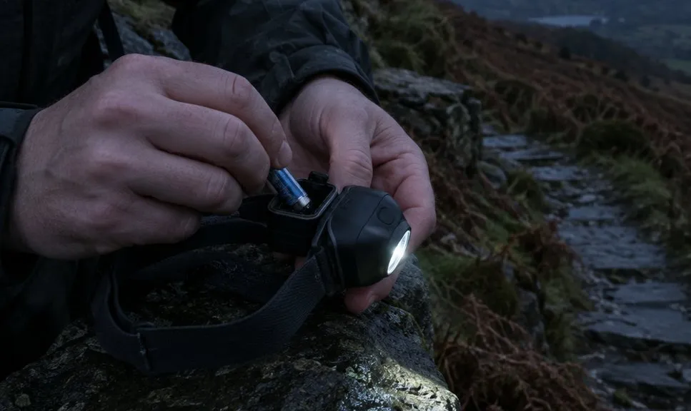 A walker's hands sliding a battery into the open compartment of a head torch, held over a wet stone wall in the last of the dusk light.