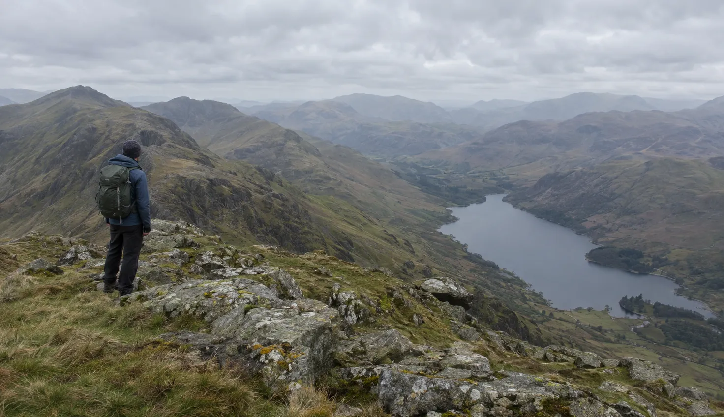 A walker on a rocky summit looking out over ridge after ridge of mountains and a long lake in the valley below.