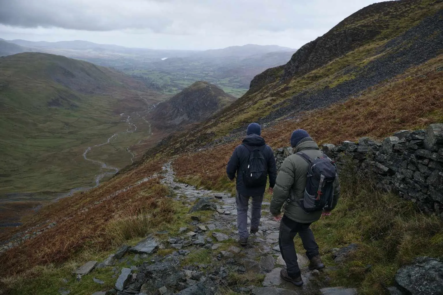 Two walkers descending a long zigzag mountain path with the valley still far below