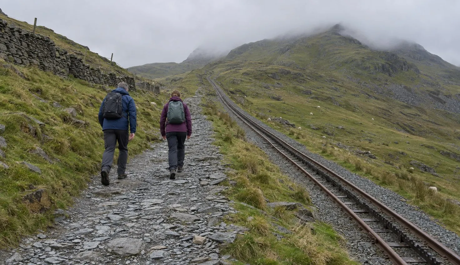 Two walkers climbing the stony Llanberis Path with the mountain railway line running beside them.