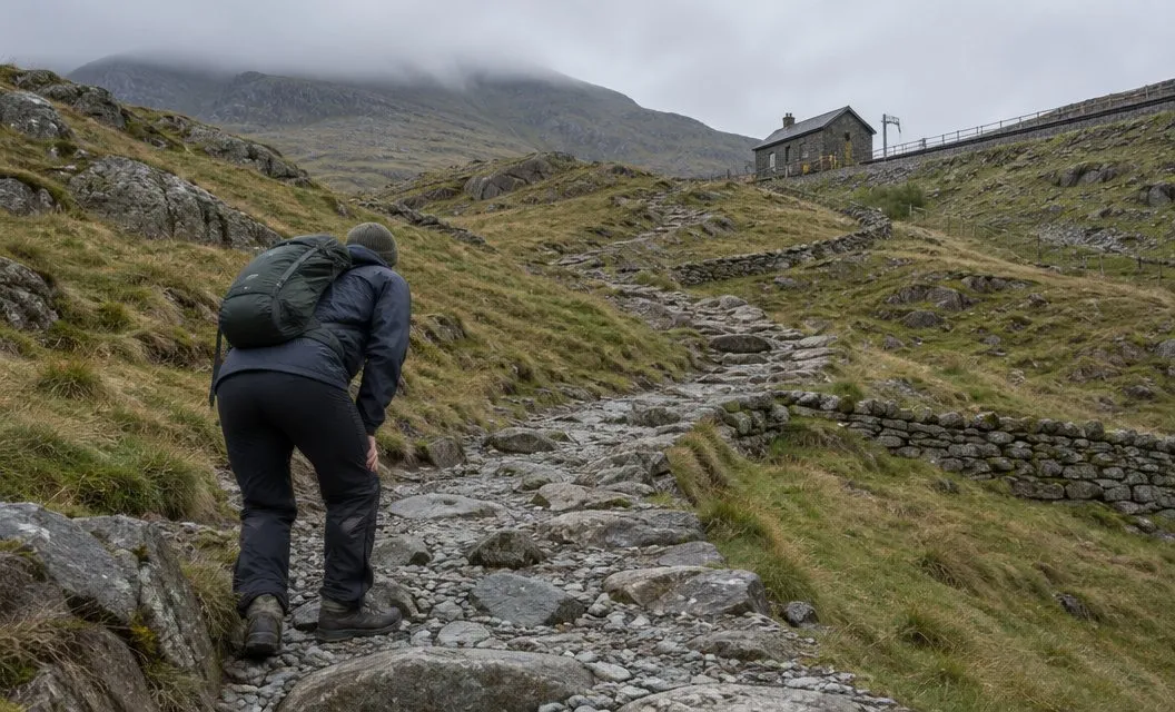 A walker pausing on a steep, stony stretch of path that climbs a bare grassy slope.