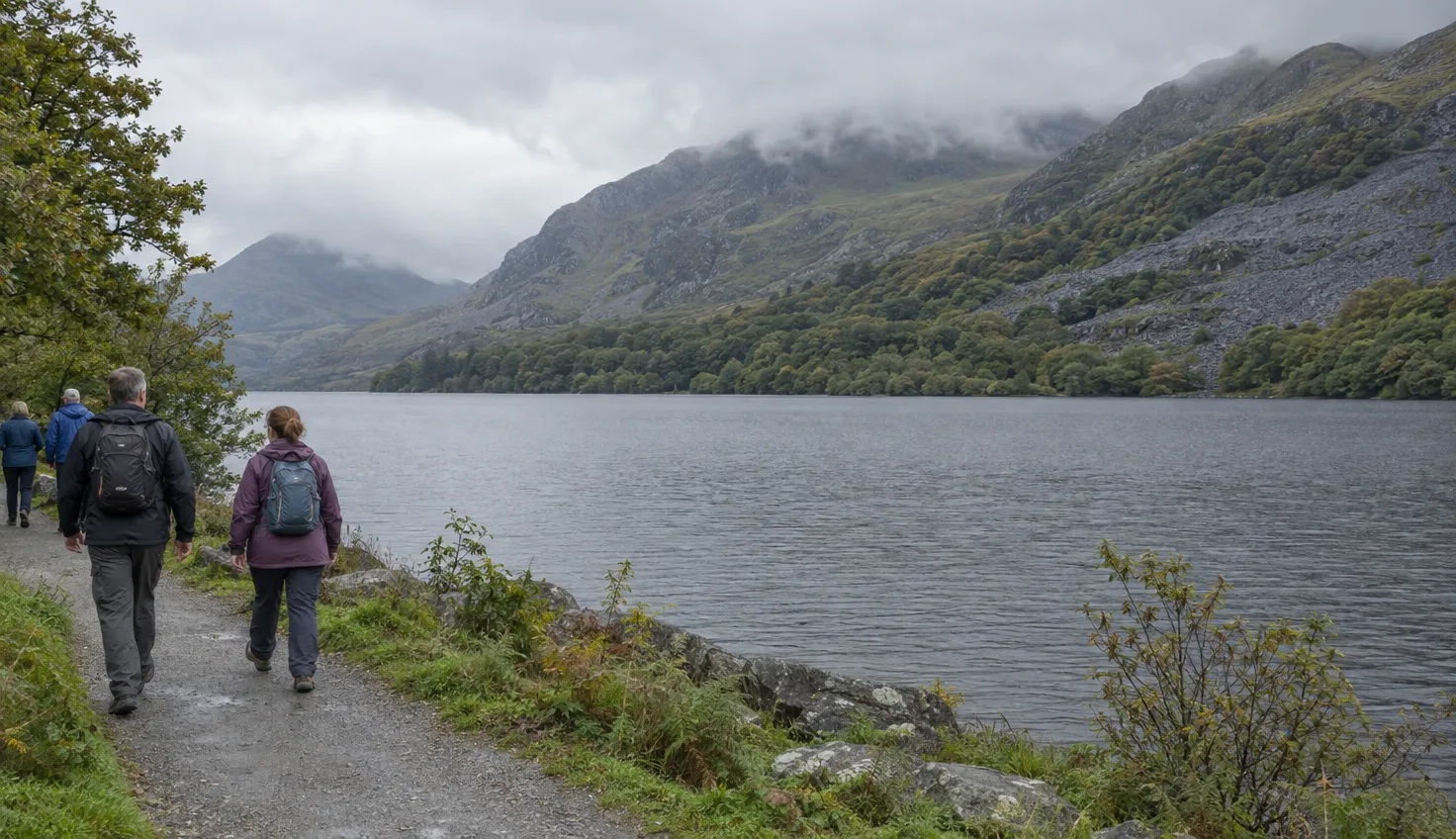 A broad Welsh lake seen from a lakeshore path, with slate slopes and oak woodland on the far bank.