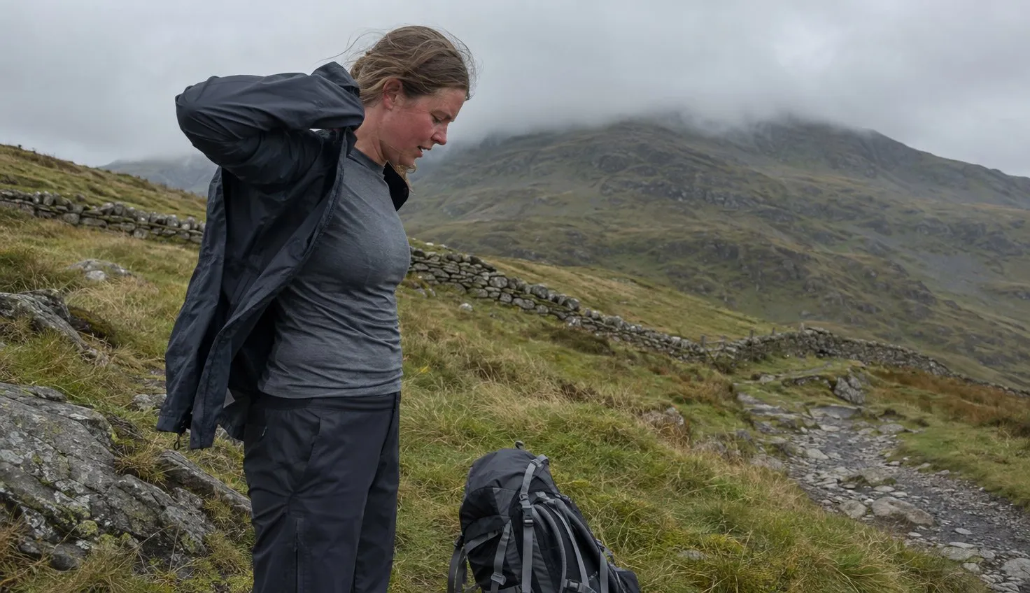 A walker at a windy col pulling a jacket back on over a thin long-sleeved base layer after the climb.