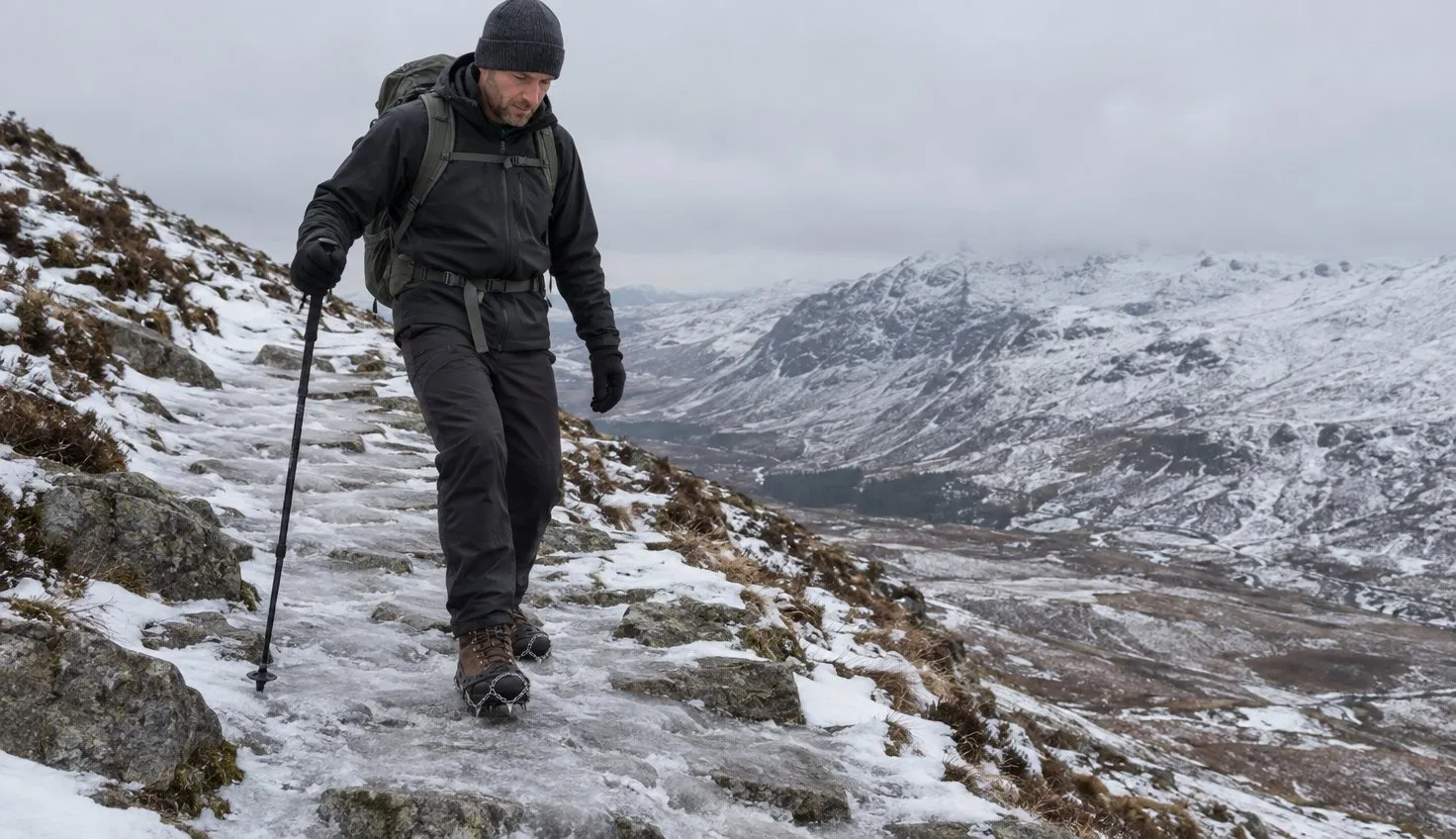 A walker in winter layers stepping down a snow-packed, icy mountain path with chain traction stretched over their walking boots.