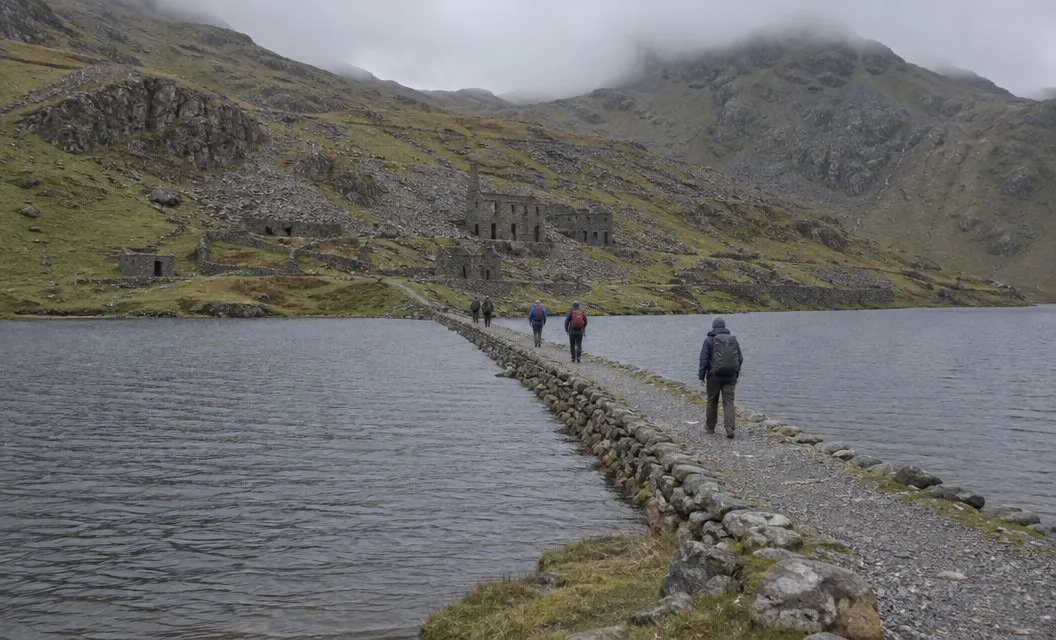 A stone causeway crossing a grey mountain lake, with walkers strung out along it.