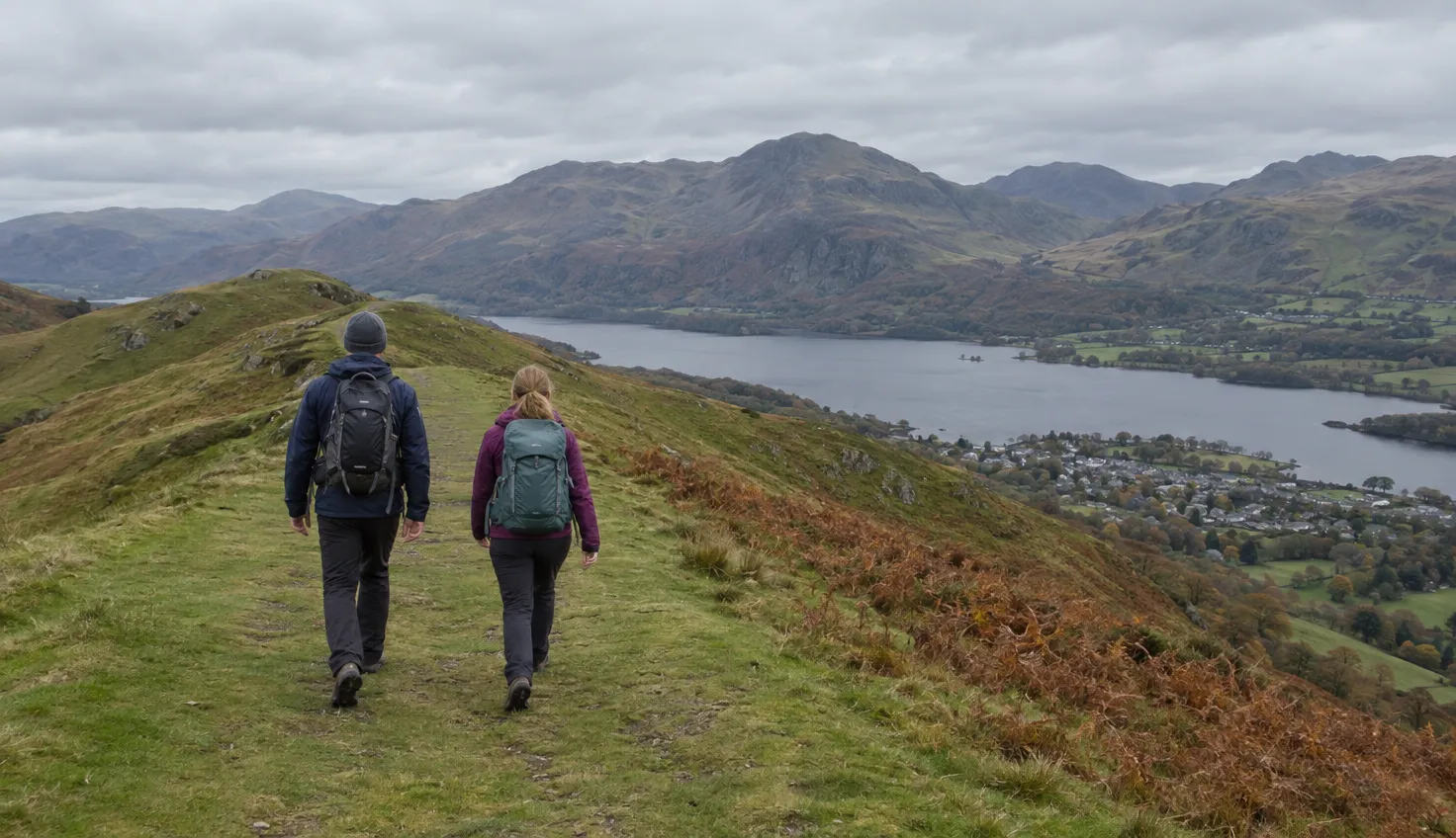 Two walkers on a broad grassy ridge, a long lake and a small town in the valley below.
