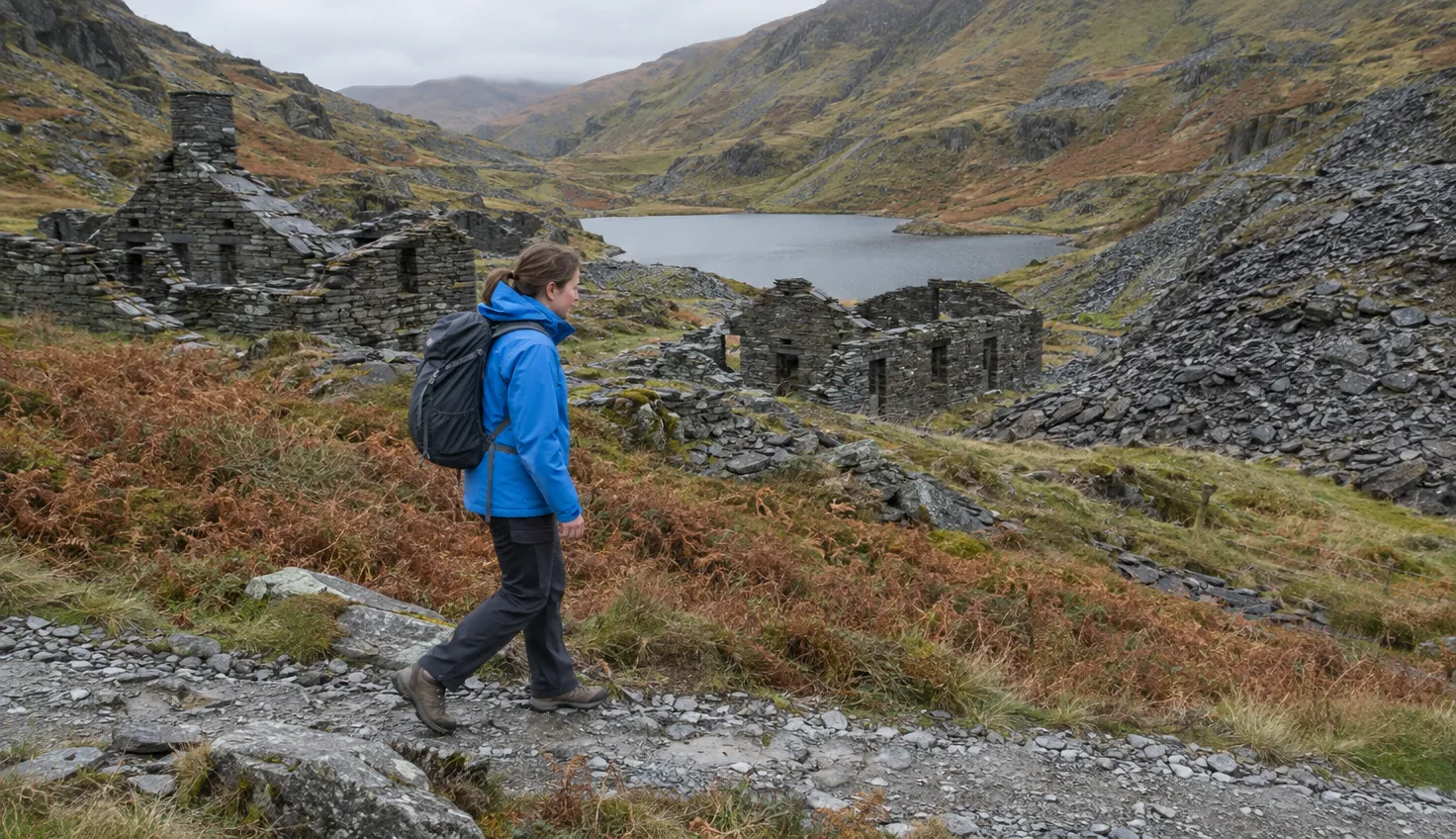 A walker on a stony path past roofless slate quarry ruins and slate tips.