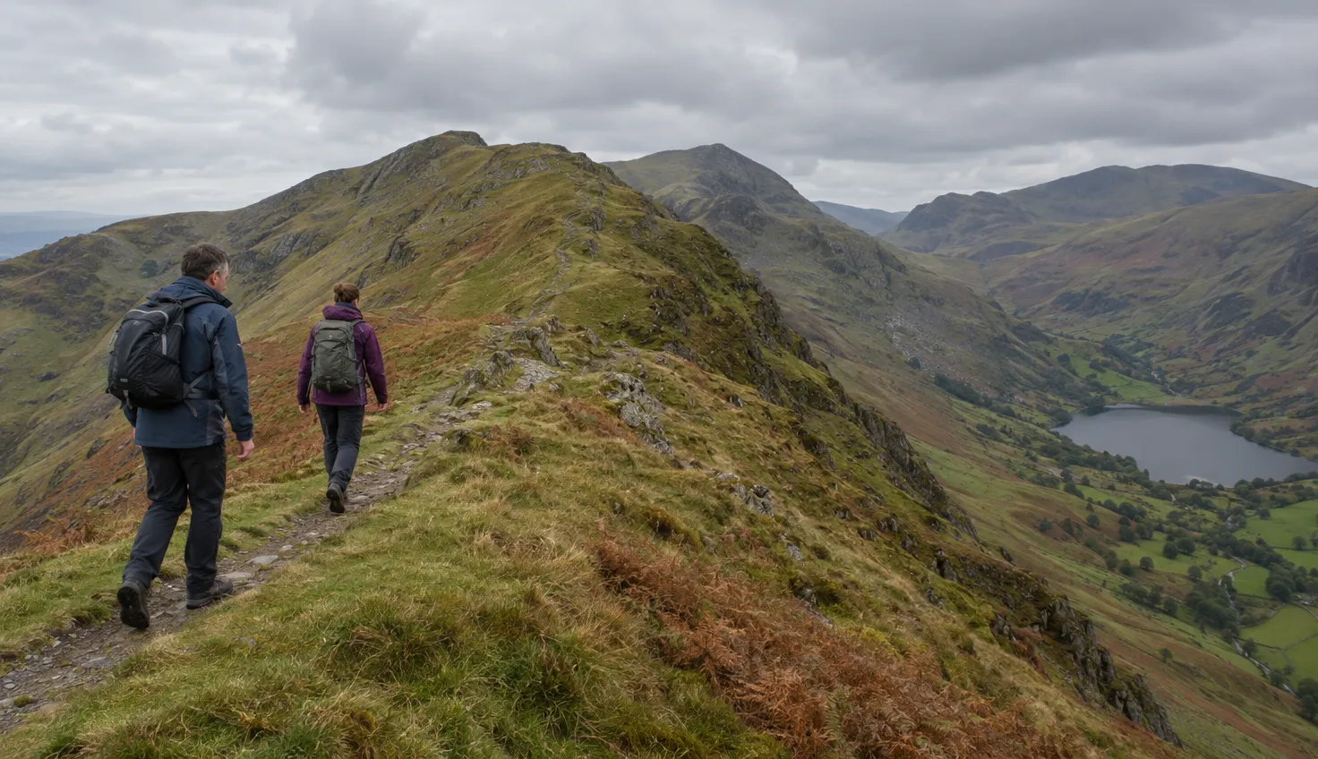 Two walkers on a narrow path along a grassy mountain ridge, a valley and small lake far below.
