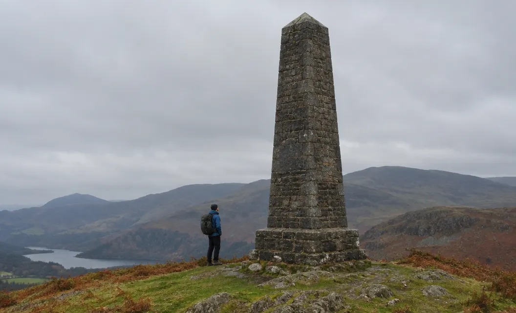 A tall stone monument on a grassy summit, a walker small beside it.