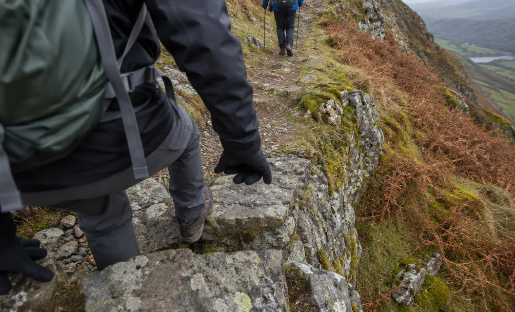 A walker's hands and boots on a short rocky step on a mountain ridge.