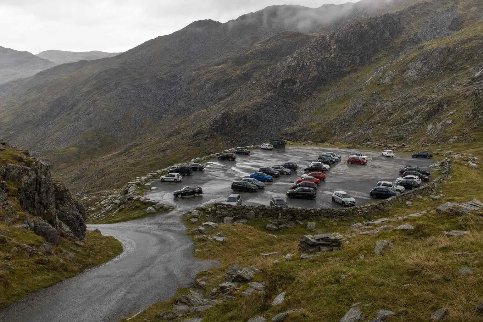 A mountain pass car park filling with cars early in the morning below slate peaks