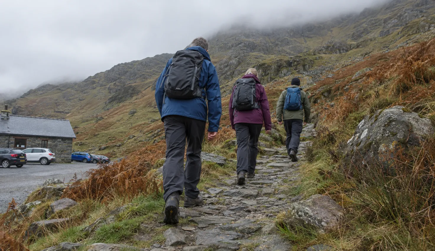 Three walkers setting off from a mountain-pass car park onto a stone path climbing into rocky hills.