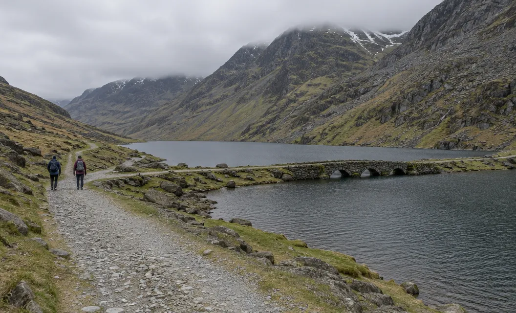 A broad stony track beside a long mountain lake with a stone causeway, two walkers small on it.