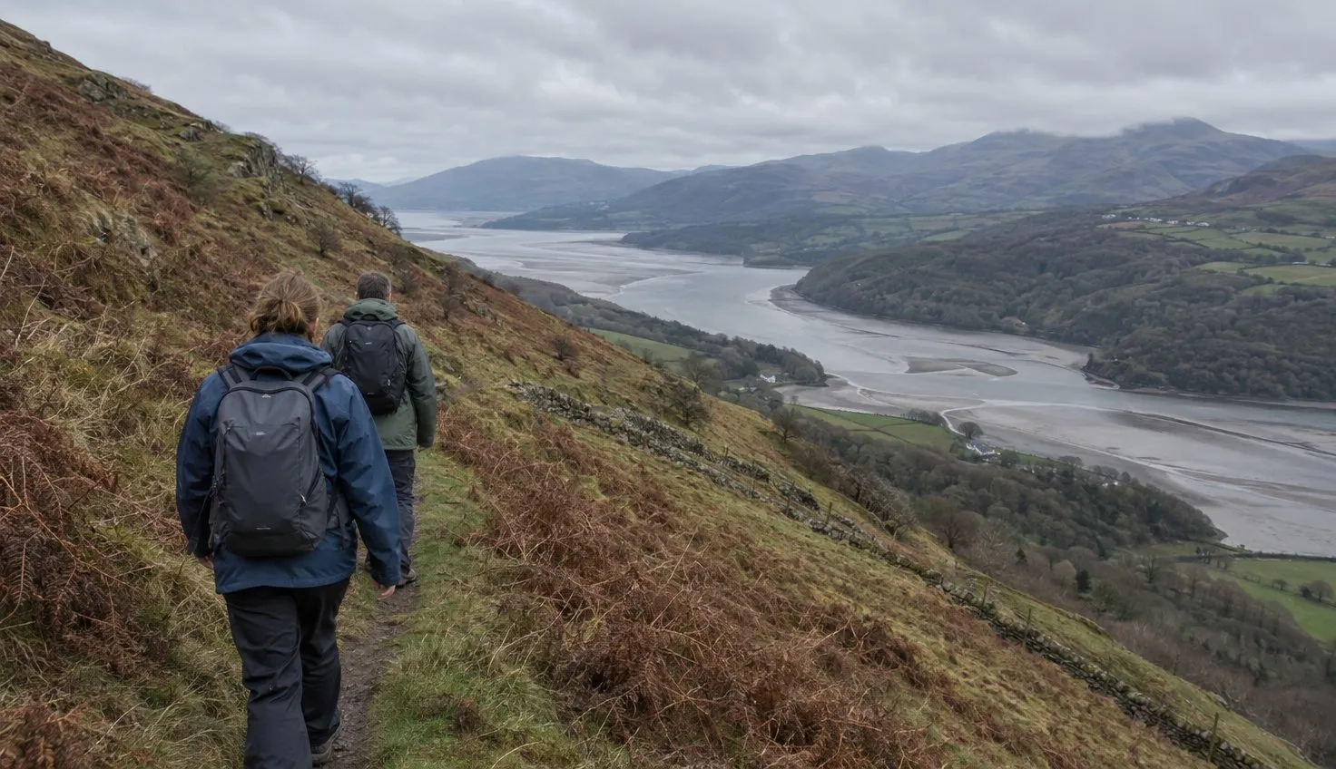 Two walkers on a narrow path across a steep hillside, high above a wide valley and river estuary.