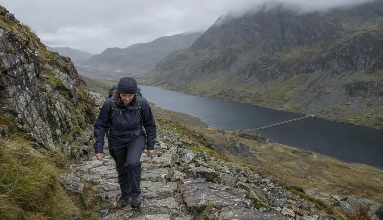 A walker stepping up rough stone steps on a rocky mountain path, with a long dark lake far below and cliffs in cloud above.
