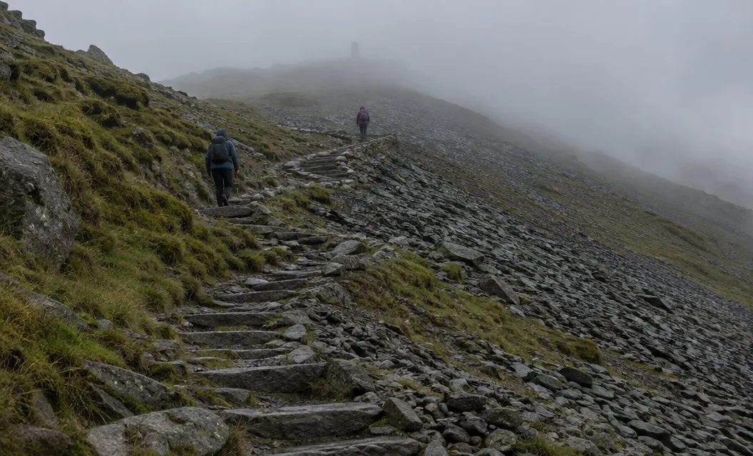 Two walkers on a steep zigzag path climbing a scree slope into thick cloud.