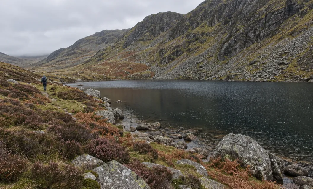 A small dark mountain lake under rocky slopes, with heather and boulders on the shore.