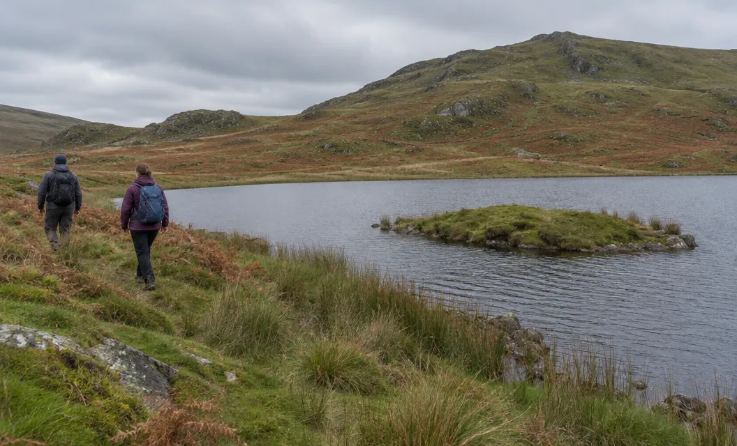 Two walkers beside a small moorland lake with a tiny grassy island near its edge.