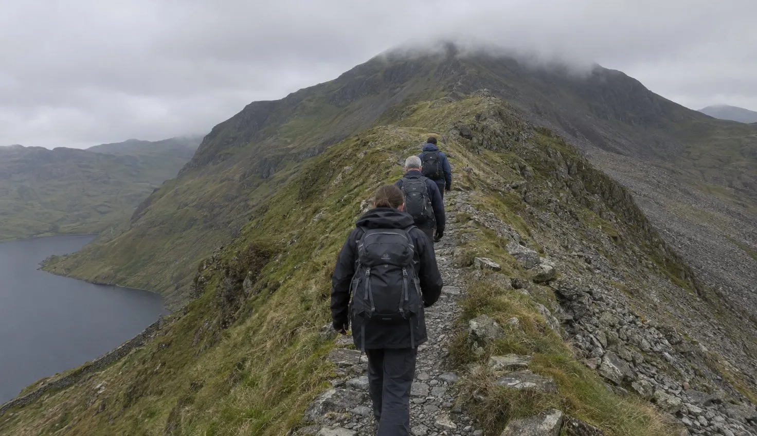 Three walkers in single file on a narrow rocky ridge path with steep slopes falling away on both sides.