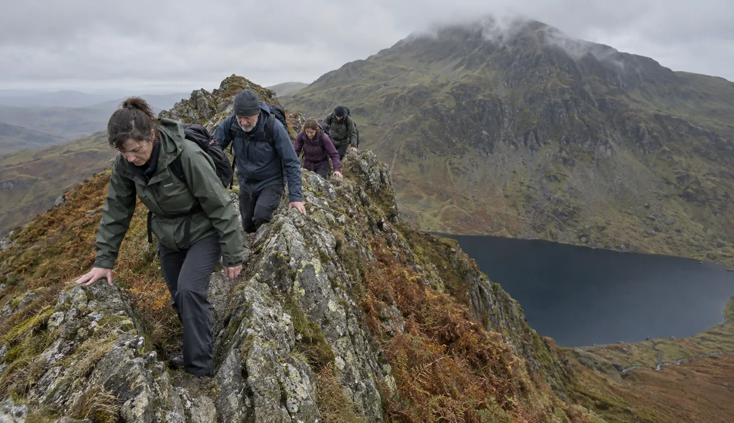 A small group of walkers in single file on a jagged rocky ridge, hands on the rock, a dark lake far below.