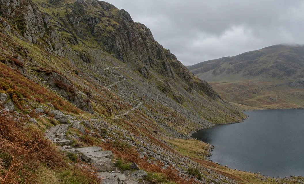A steep rocky mountain face with a faint path through scree and rock steps above a lake.