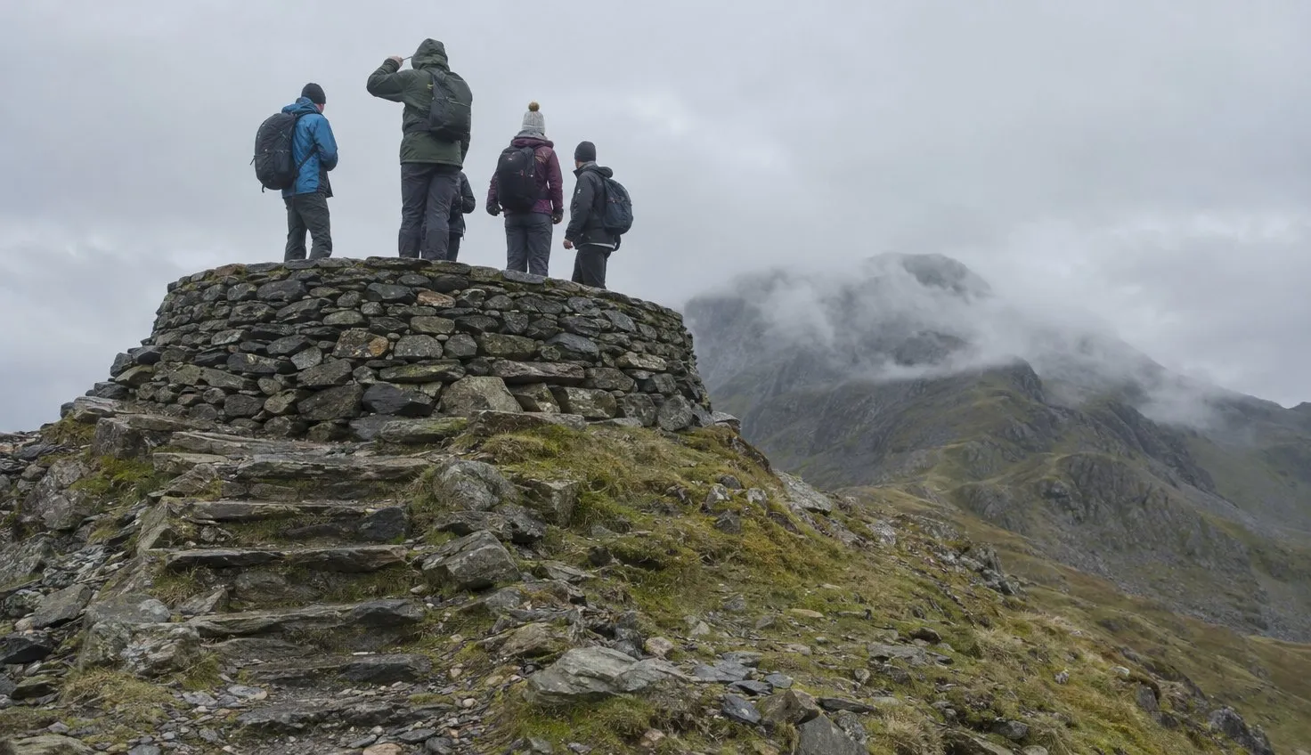 Walkers gathered on a rocky summit cairn with stone steps, cloud drifting across the ridge below.