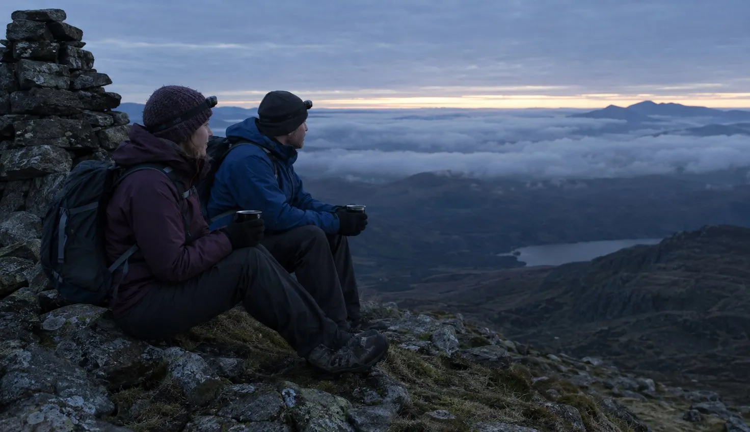 Two walkers huddled by a summit cairn in warm layers, watching a pale band of first light above the cloud.