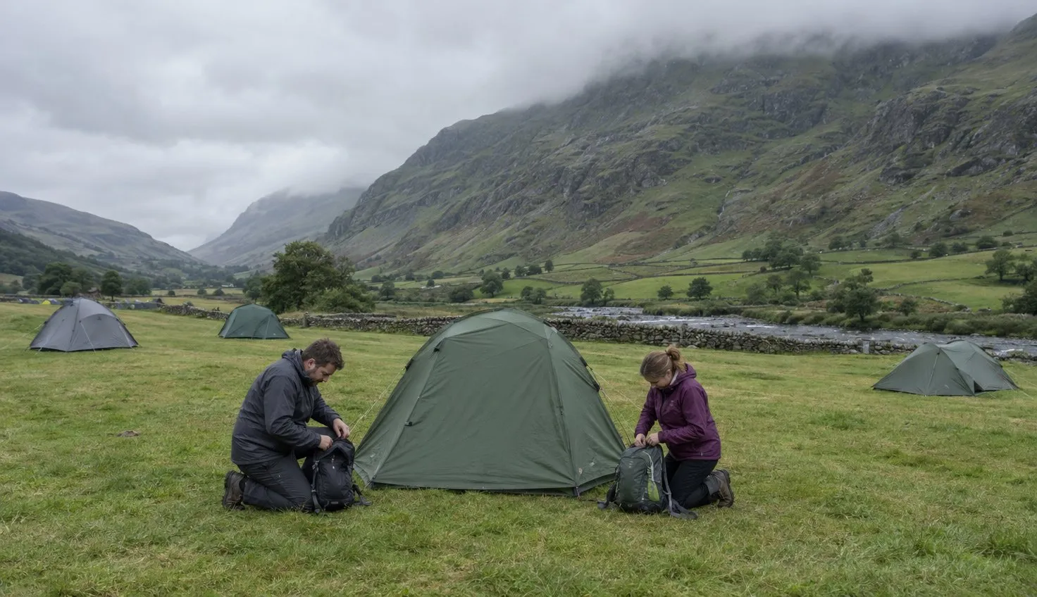 Two walkers packing daypacks beside their tent on a campsite field below a mountainside.