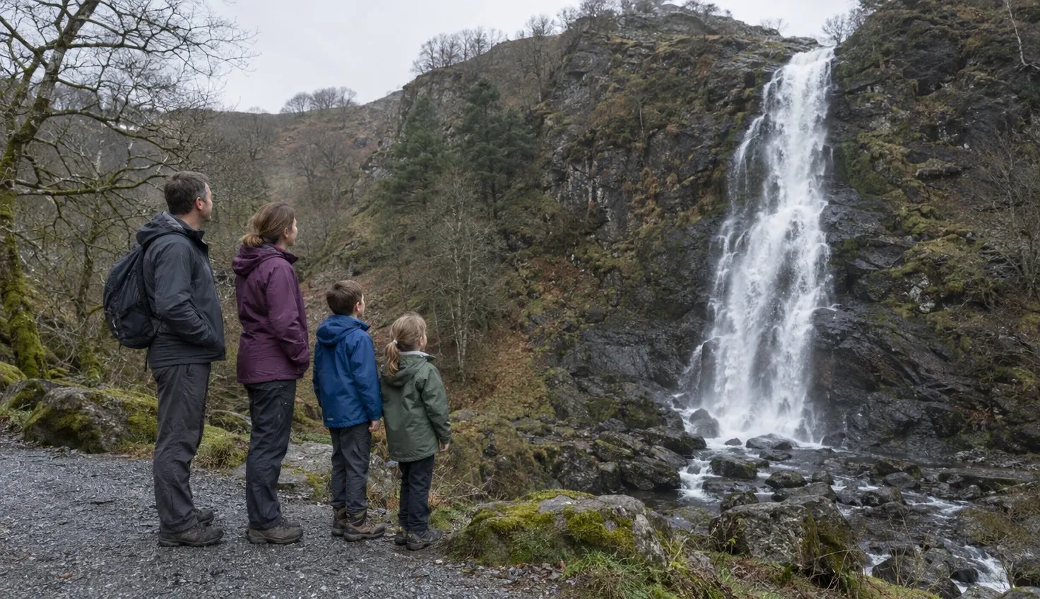 A family in waterproofs on a valley path, looking up at a tall waterfall on a dark rock face.