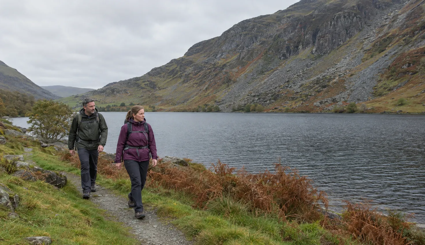 Two walkers on a grassy path beside a long lake, a steep mountainside rising beyond.