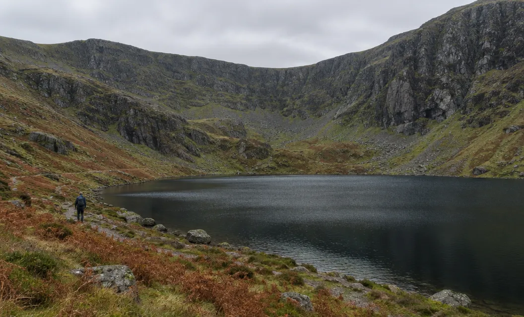 A dark corrie lake below a huge curving wall of cliffs, a walker small on the shore.