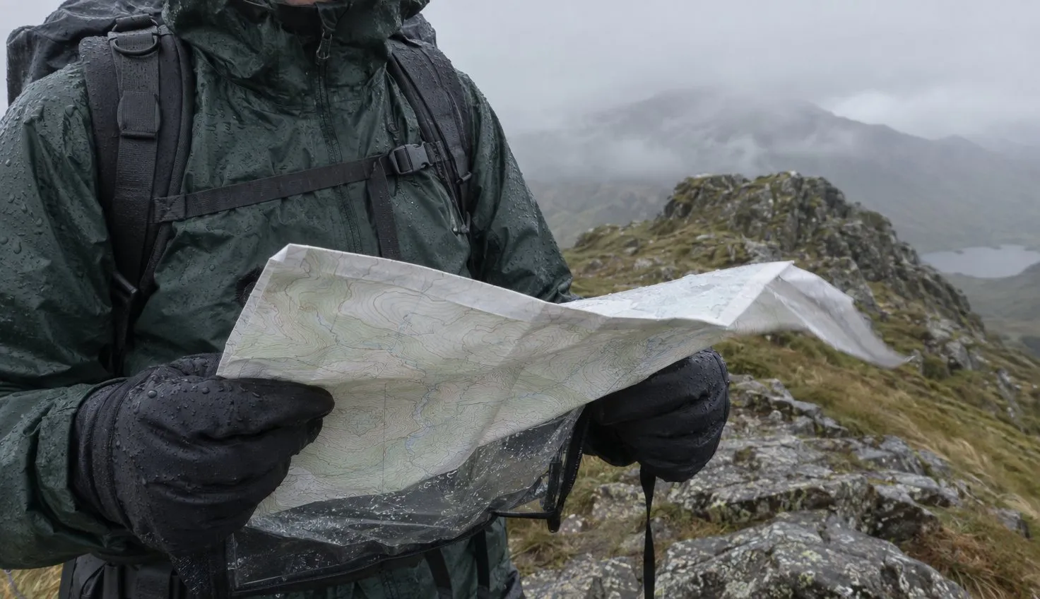A walker's gloved hands holding a folded map in the wind on a wet, misty summit ridge.