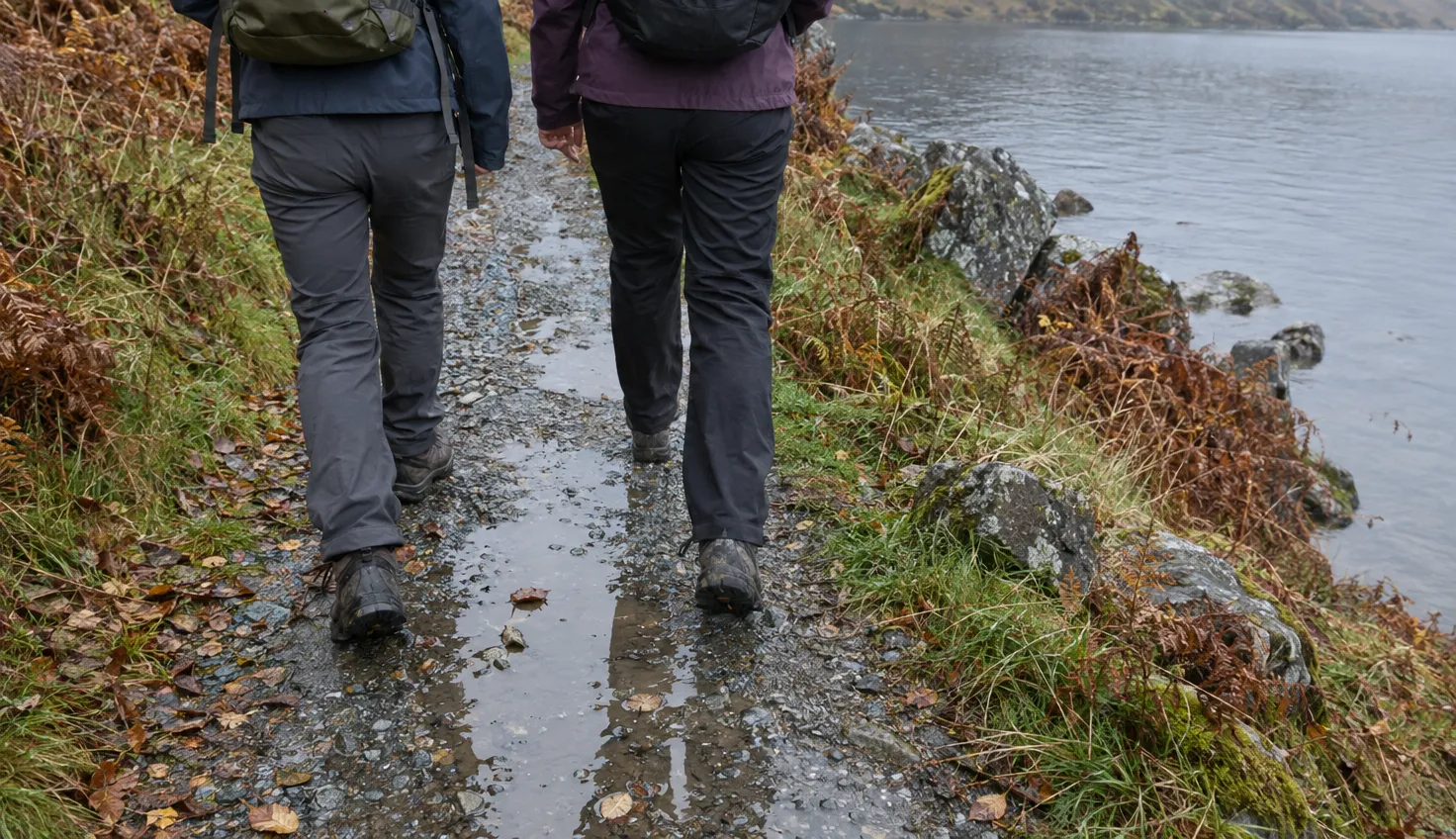 Two walkers in low walking shoes on a wet gravel path beside a lake.