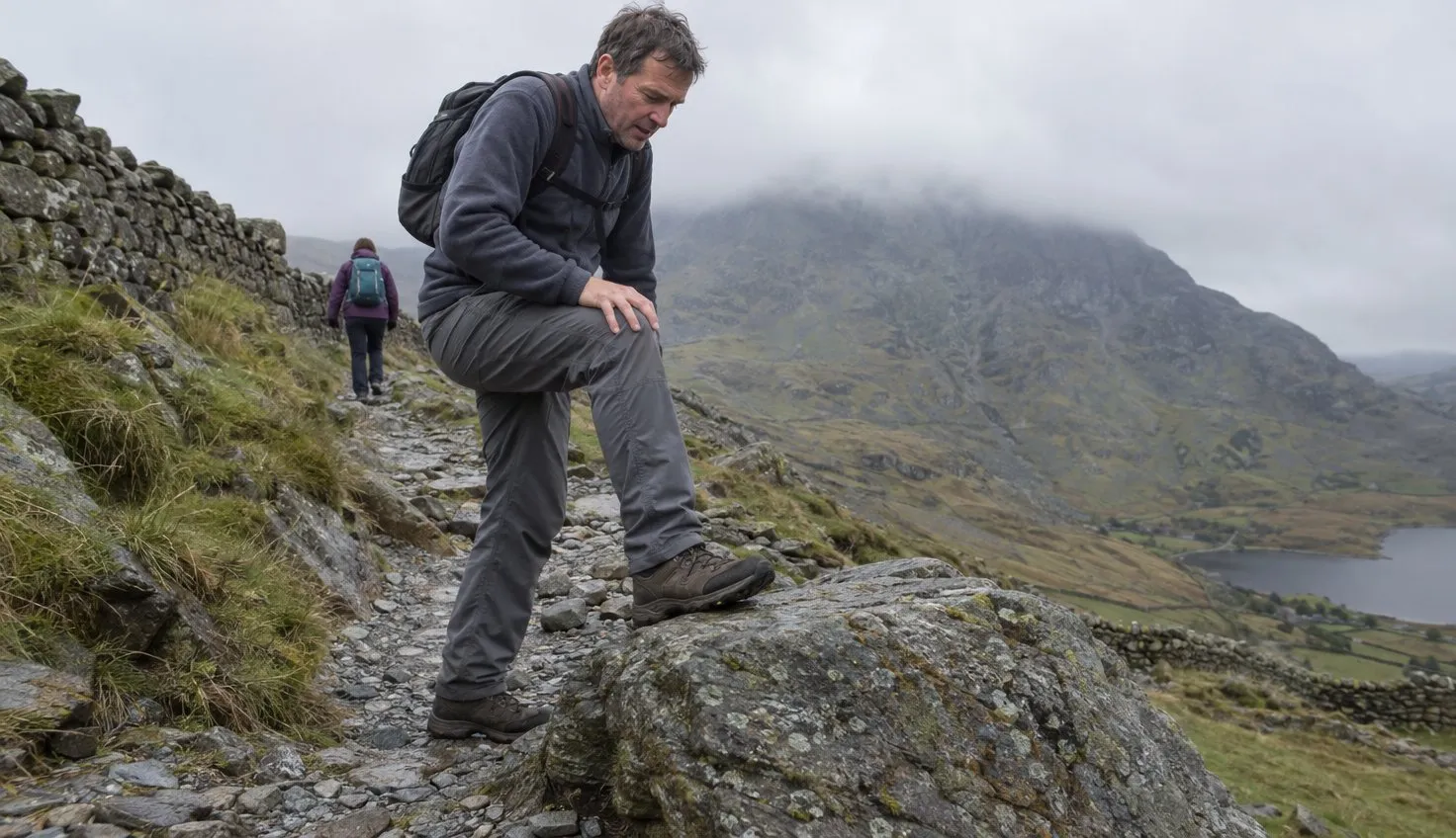 A walker in grey walking trousers taking a big step up onto a rock on a stony mountain path.