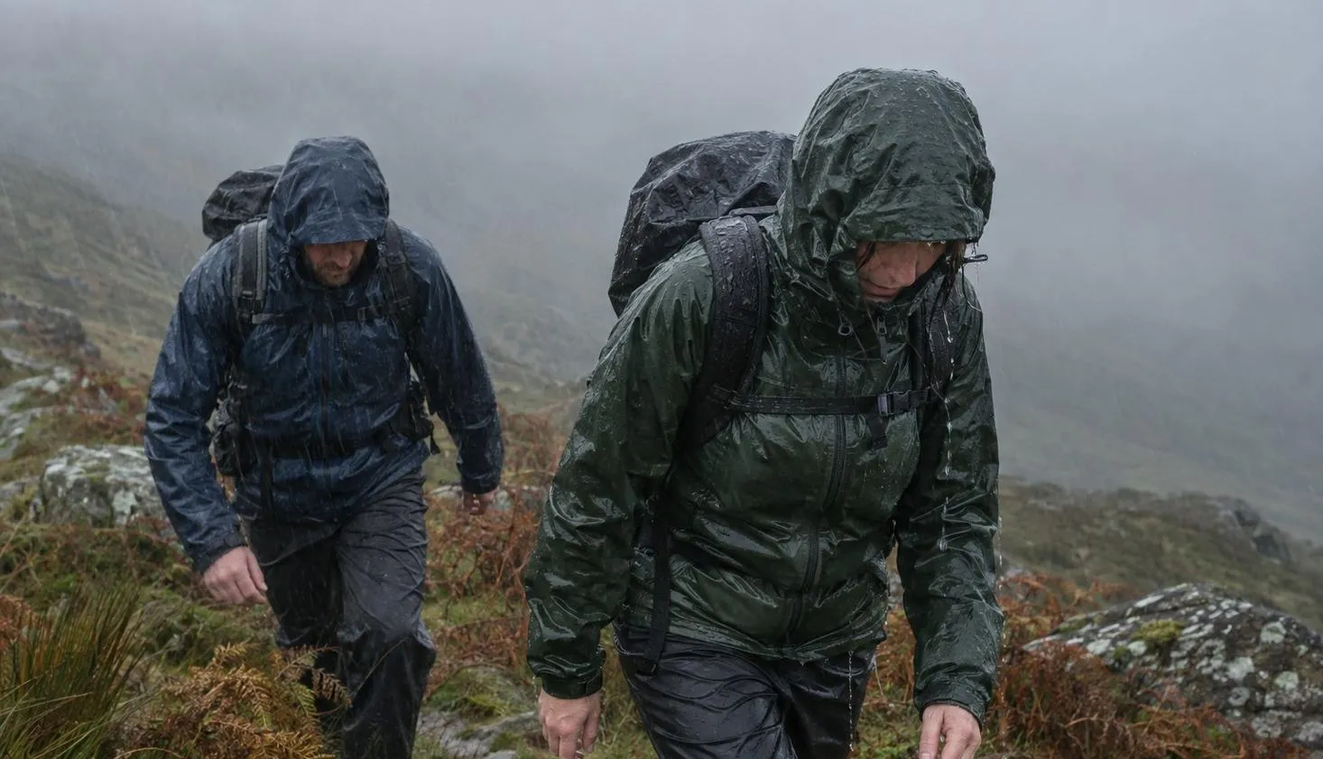 Two walkers in soaked waterproof jackets with hoods up, heads down in driving rain on an open Welsh hillside.
