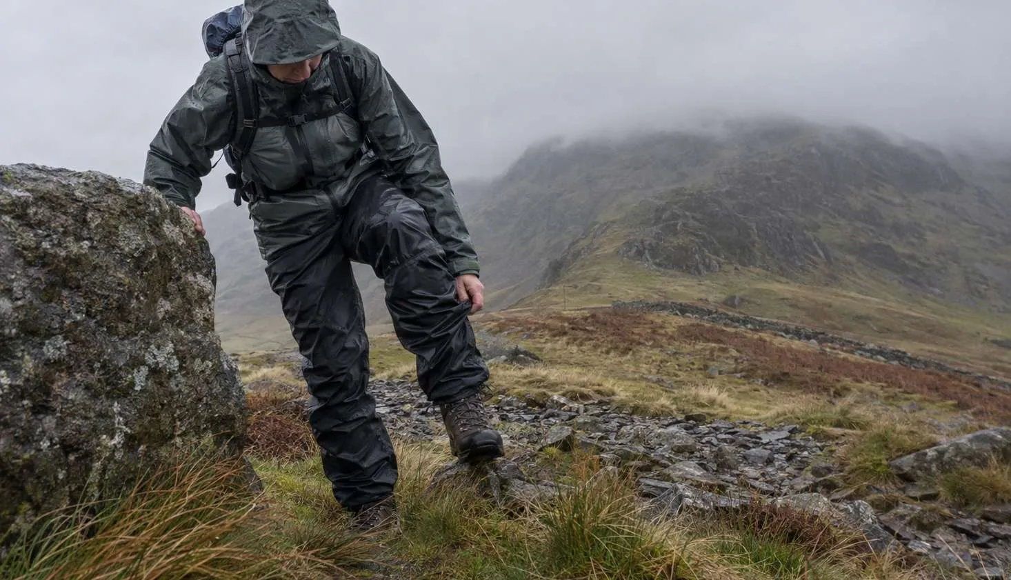 A walker on a windswept Welsh col, braced against a boulder, pulling waterproof overtrousers up over a laced walking boot while the wind pushes the fabric sideways.
