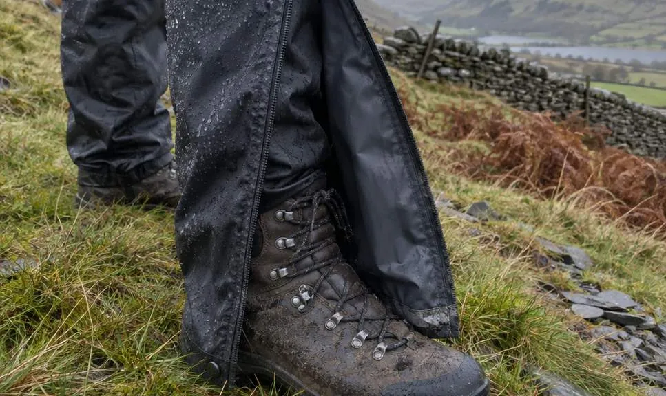A waterproof overtrouser leg opened along a long side zip and folded back over a still-laced walking boot on wet hillside grass.